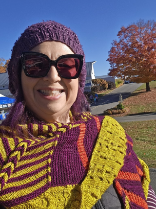 white woman smiling at camera with purple/yellow/orange scarf, purple hair, and purple knit cap