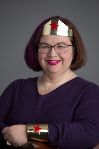 white woman smiling at camera with purple shirt, glasses, and wonder woman crown and arm band